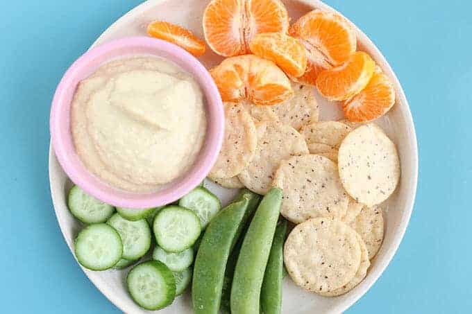 homemade hummus without tahini in pink bowl with cucumbers, snap peas, crackers on plate