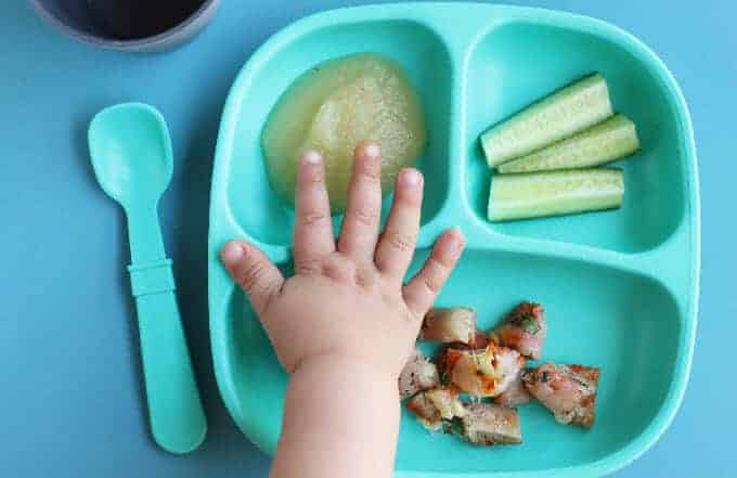 toddler dinner plate with pizza, cucumbers, and applesauce and toddler hand