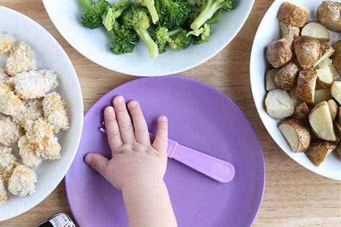 family style meal with chicken nuggets, broccoli, potatoes, and toddler hand over purple plate