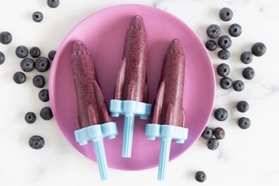 Blueberry popsicles on purple plate with blueberries on side.