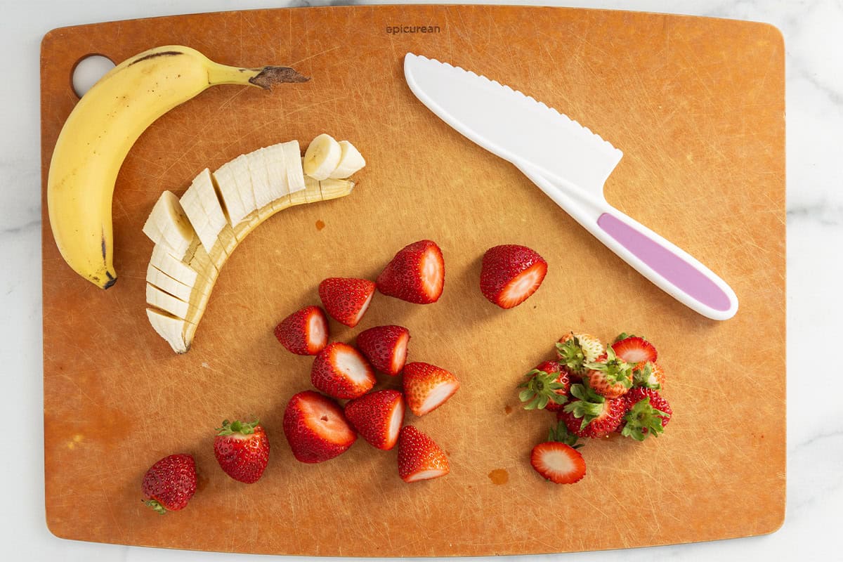 Strawberries and banana being chopped on cutting board for santas.