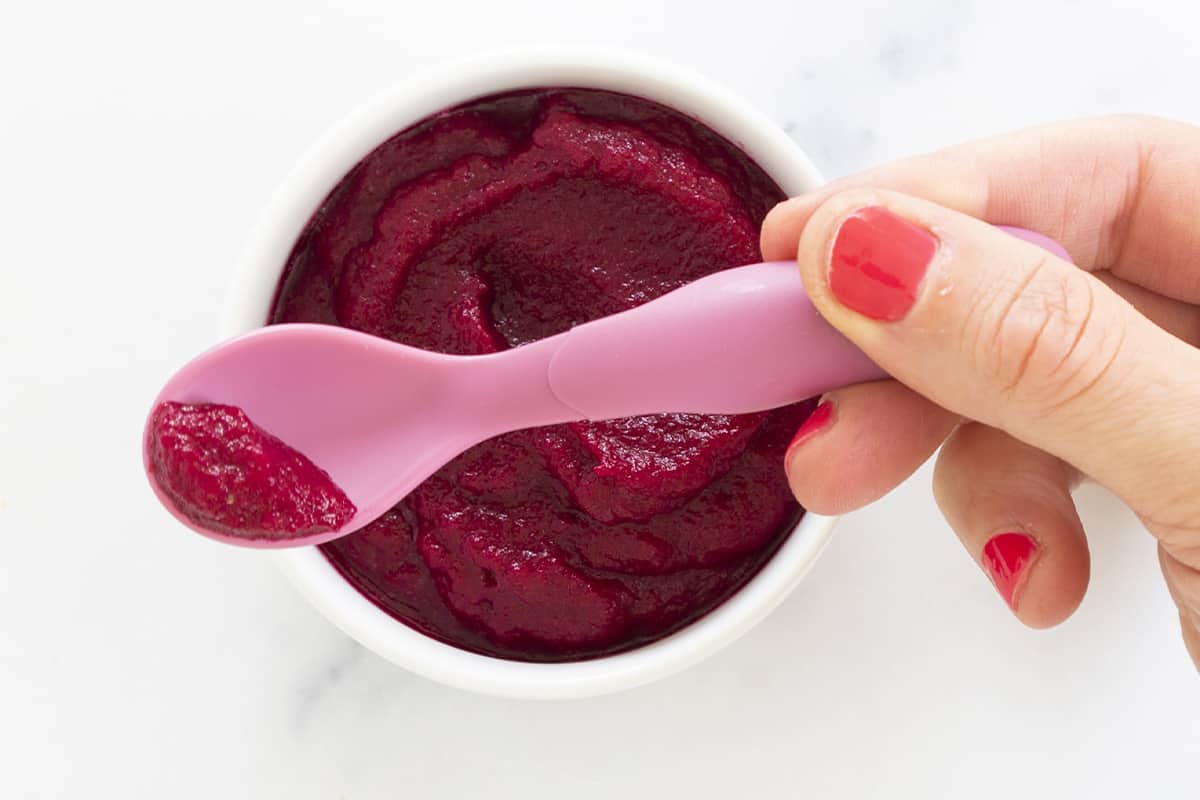 beets baby food in bowl with hand holding pink spoon over top.