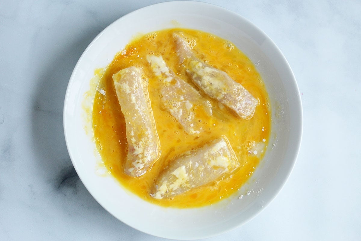 fish strips coated in egg in white bowl.