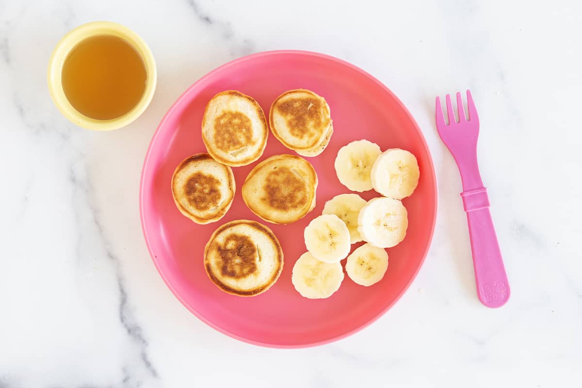 Mini pancakes on kids pink plate with bananas.