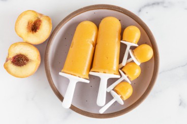 Peach popsicles on grey plate with peach on side.