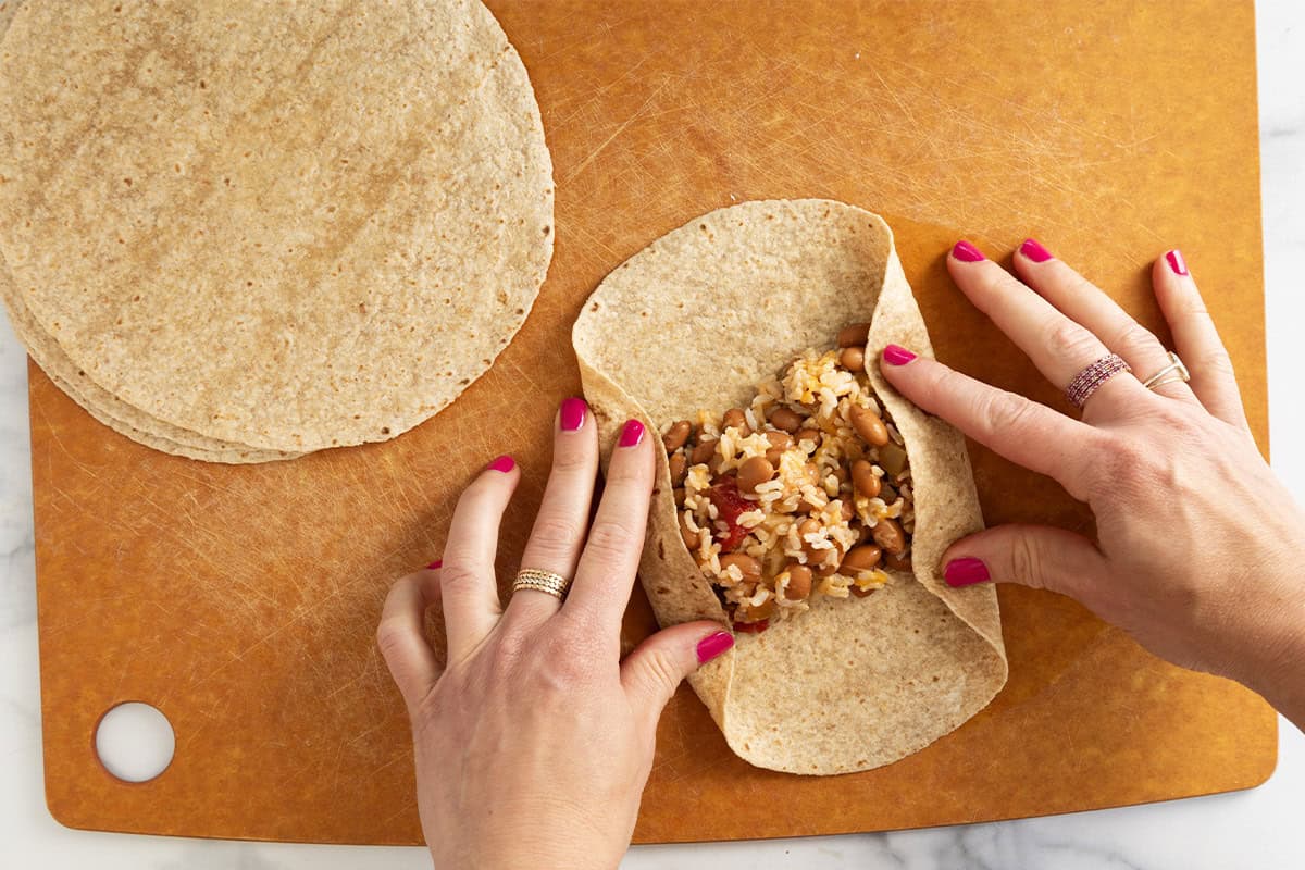 Hands wrapping up rice and bean burritos on cutting board.