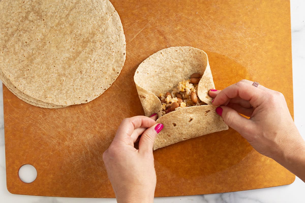 Hands wrapping up rice and bean burritos on cutting board.