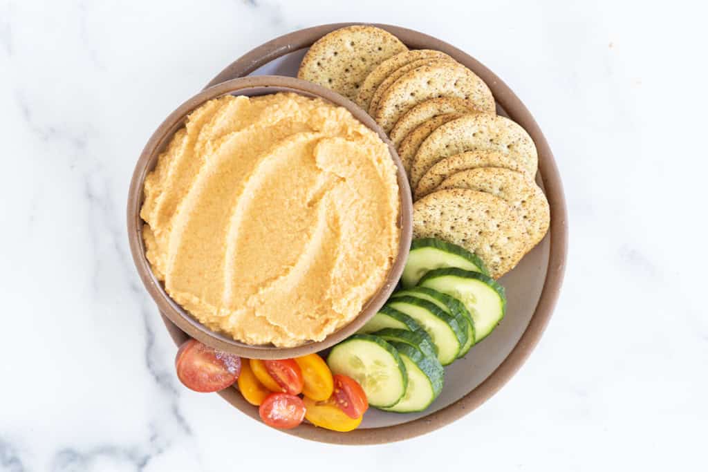 Carrot hummus in bowl with crackers and veggies on side.