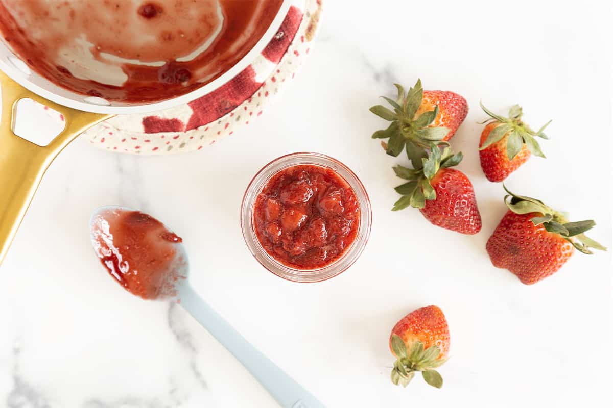 Strawberry sauce in glass jar with pan and spoon.
