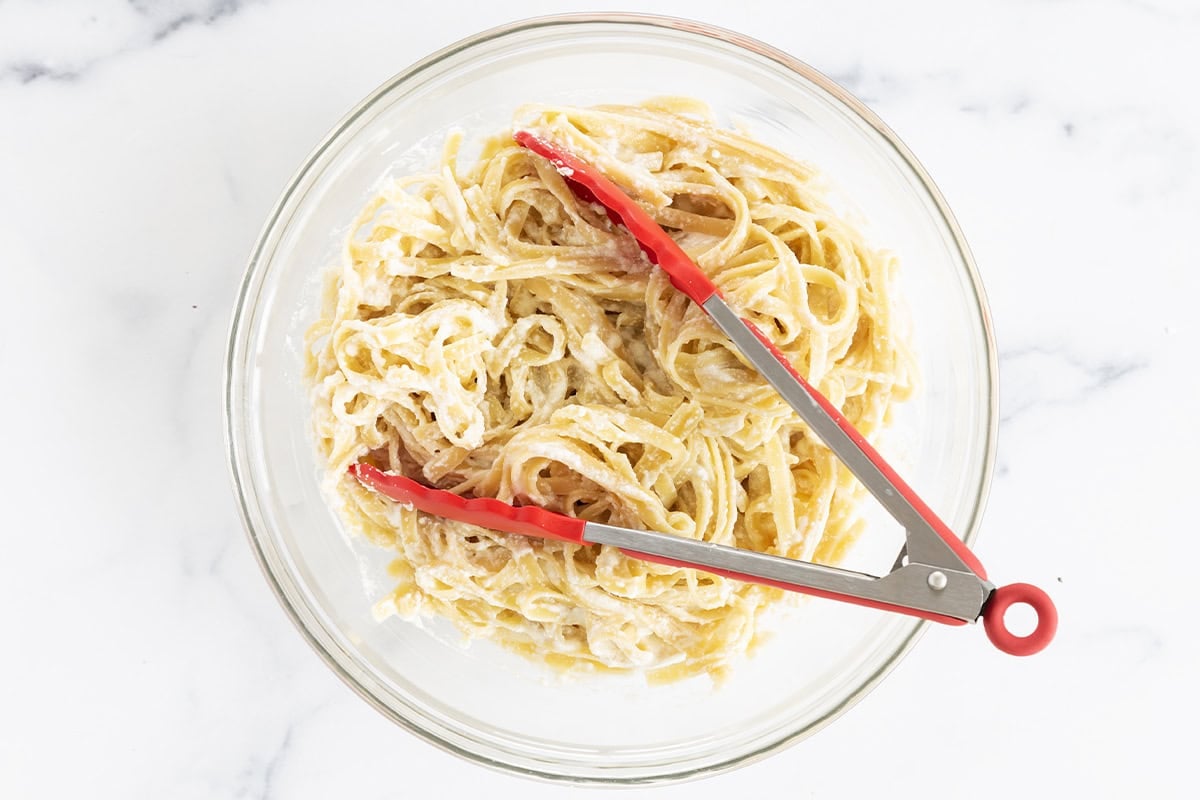 Mixing together lemon ricotta pasta in glass bowl with thongs.