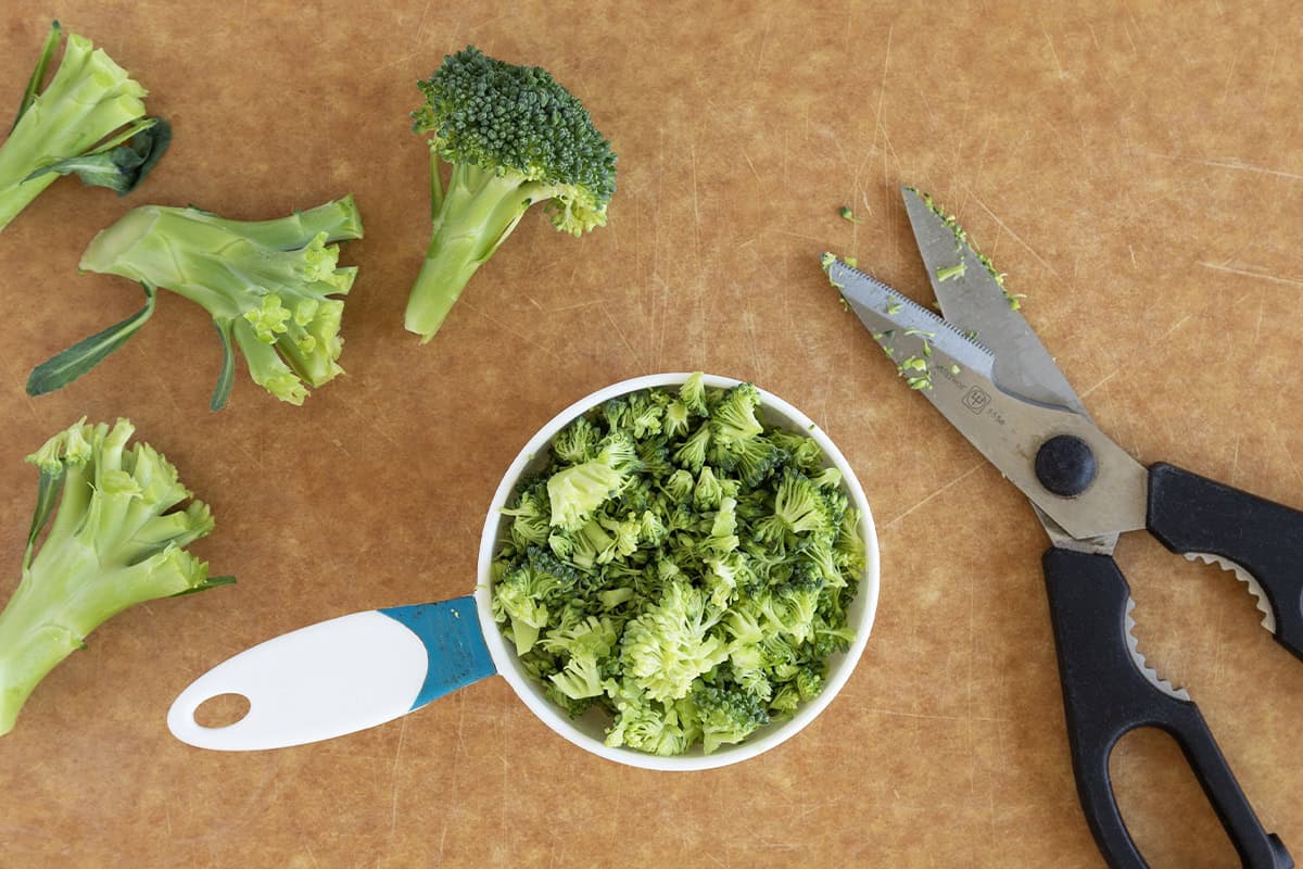 Broccoli on cutting board with scissors and measuring cup for an egg bake 