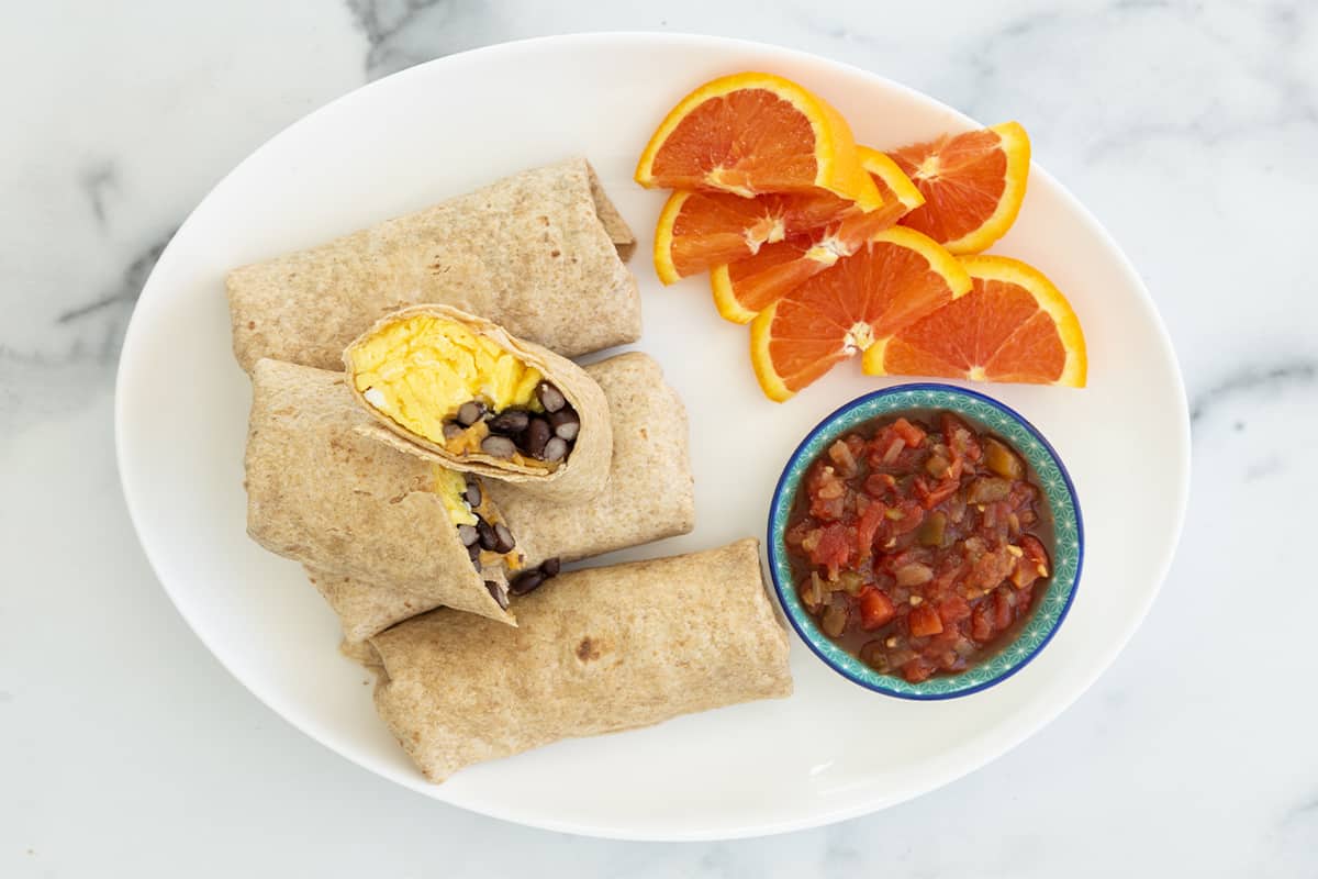 breakfast burritos, one cut in half, on plate with oranges and salsa on side.