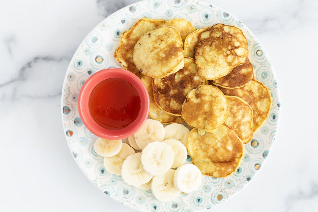 2-Ingredient banana pancakes on plate with banana slices and syrup.