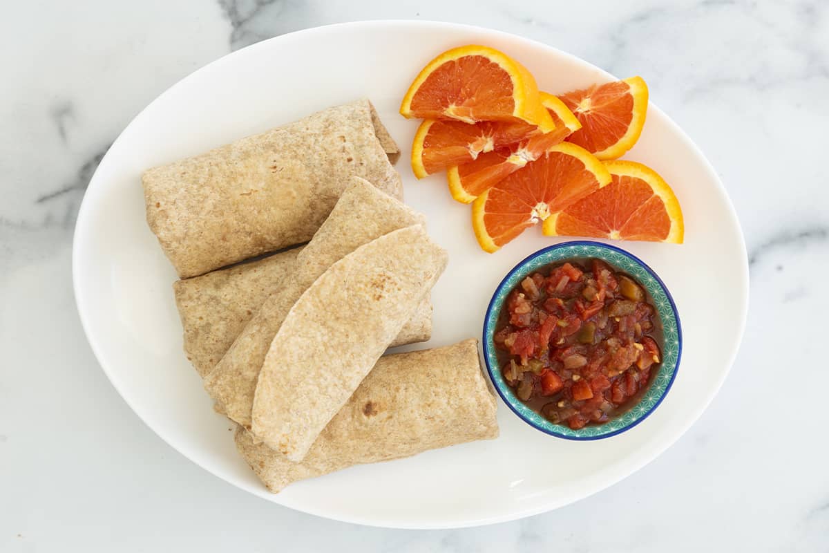 wrapped breakfast burritos on serving platter with sliced oranges and salsa on the side.