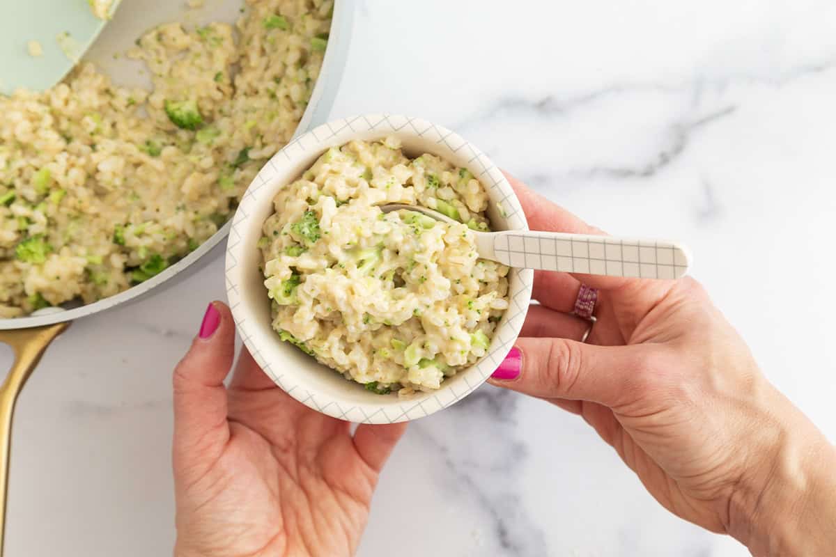 Hands holding cheesy broccoli rice in white bowl.