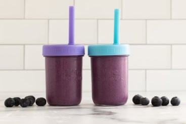 Blueberry smoothie in two cups with lids and straws.