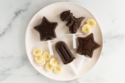 Chocolate banana popsicles on white plate with banana slices.