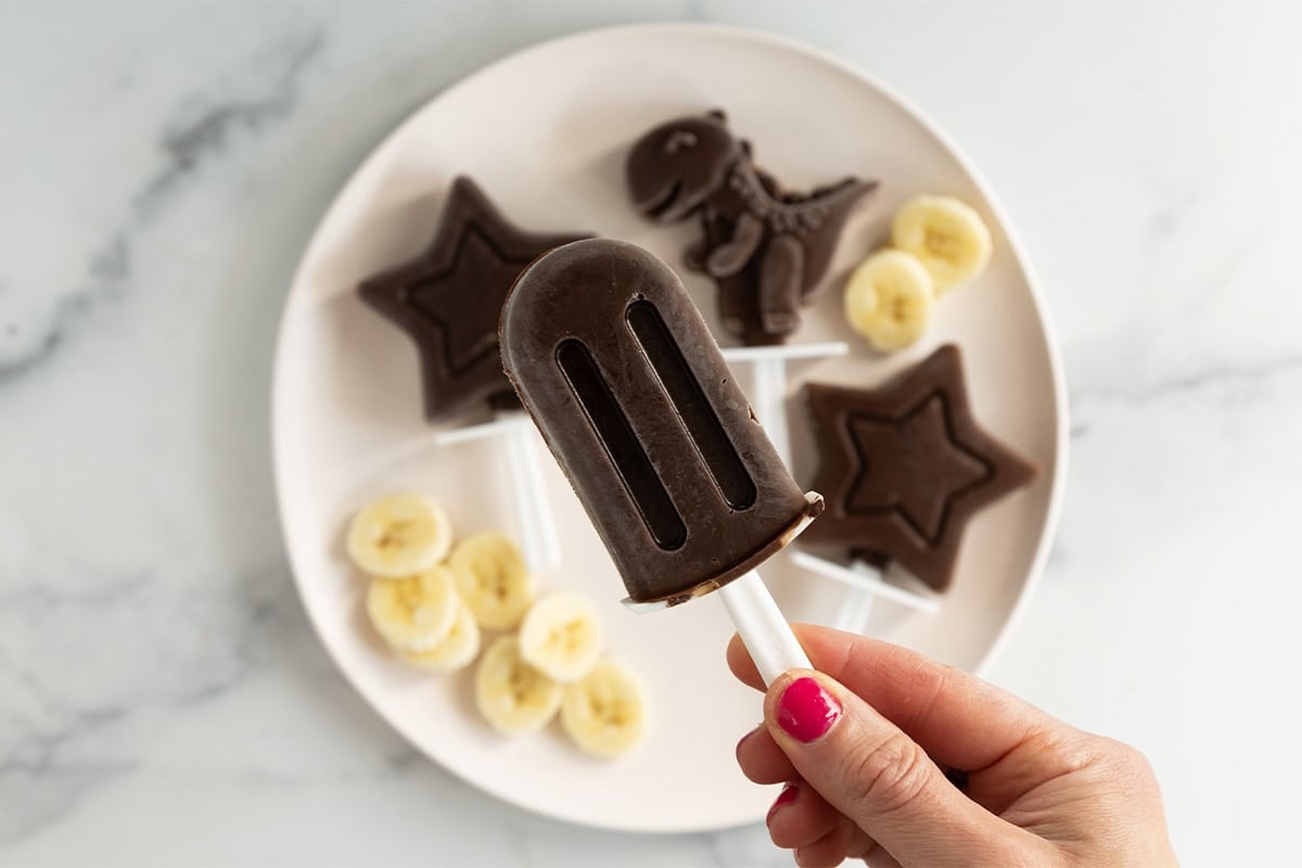 Hand holding Chocolate banana popsicles over white plate with banana slices.