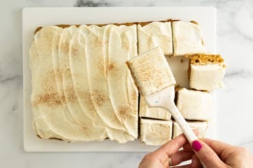 Pumpkin bars with frosting, hands holding bar on spatula.