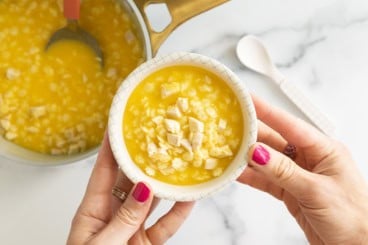 Hands holding white bowl of baby soup over pot.