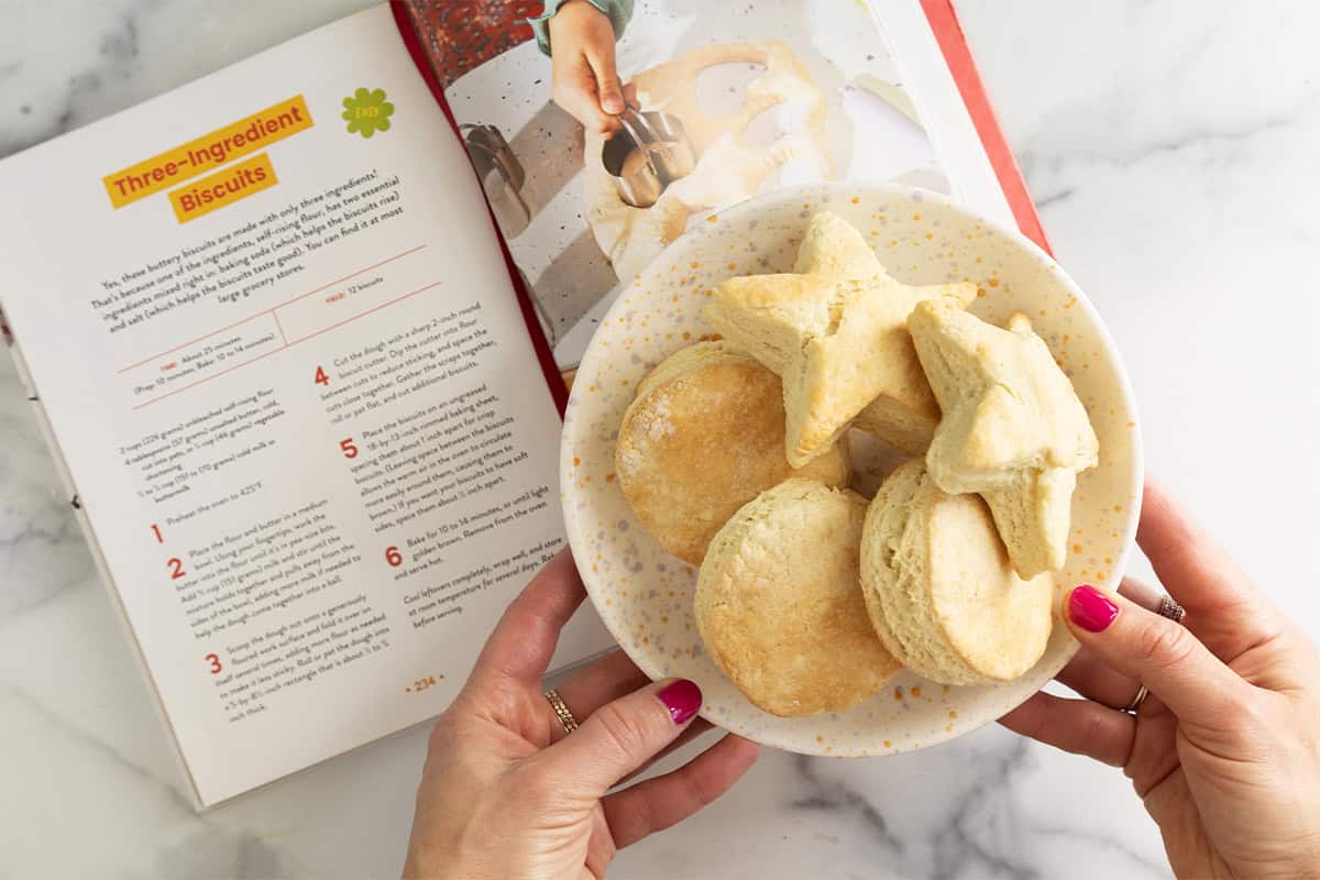 Hands holding plate of biscuits over cookbook.