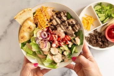 Hands holding white bowl of cheeseburger salad.