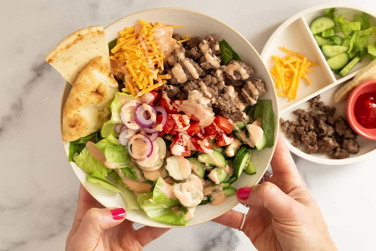 Hands holding white bowl of cheeseburger salad.