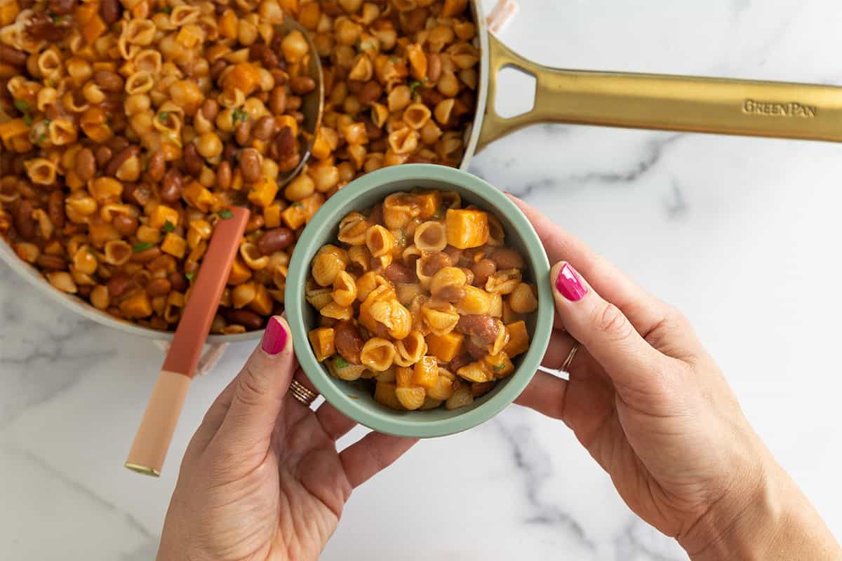 Hands holding bowl of veggie chili mac over top of pan.