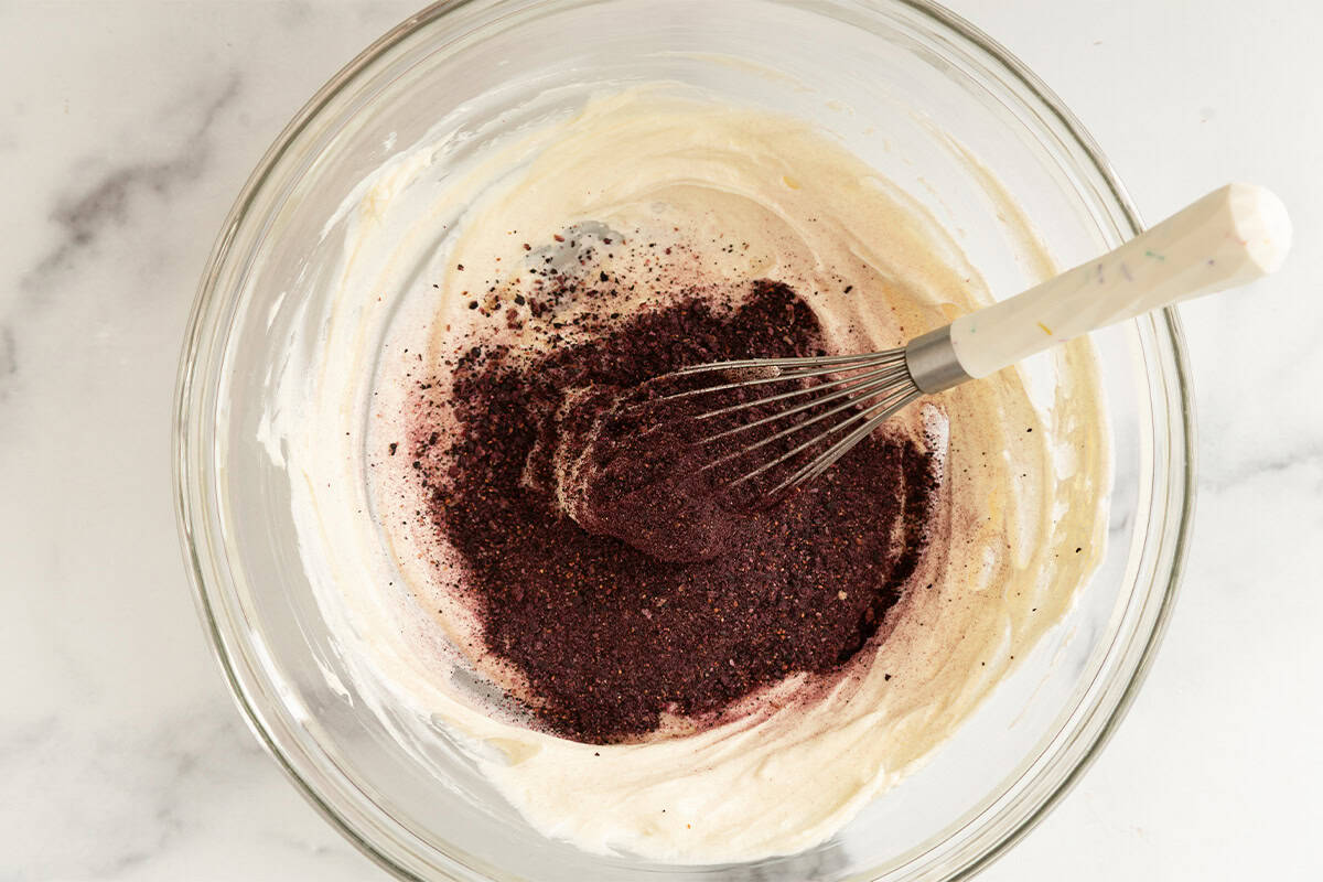 Adding ground up blueberries to cream cheese frosting in glass bowl.