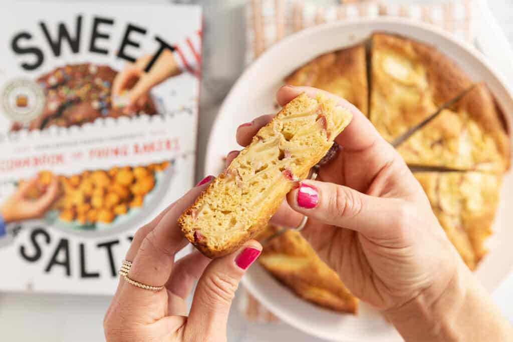 slice of apple cake in hand with cookbook in background.
