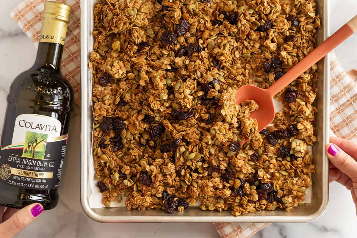 Olive oil granola on baking sheet with spatula and olive oil bottle on side.