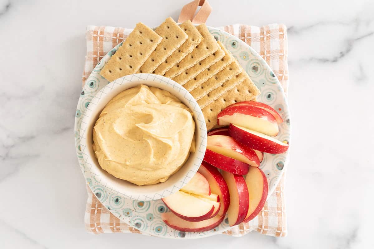 Pumpkin cheesecake dip in bowl with apples and crackers on side.