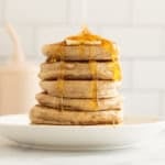 Stack of pancakes without eggs on white plate with syrup being poured on top.