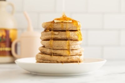 Stack of pancakes without eggs on white plate with syrup being poured on top.