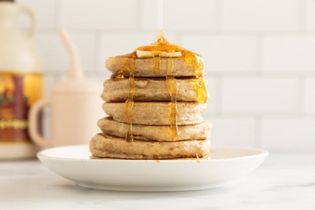 Stack of pancakes without eggs on white plate with syrup being poured on top.