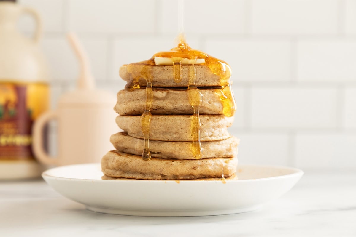 Stack of pancakes without eggs on white plate with syrup being poured on top.