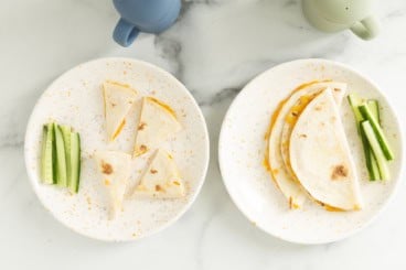 Cheese quesadillas on two white plates with cucumber sticks.