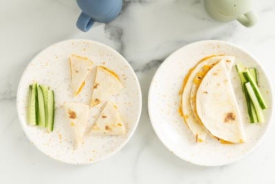 Cheese quesadillas on two white plates with cucumber sticks.
