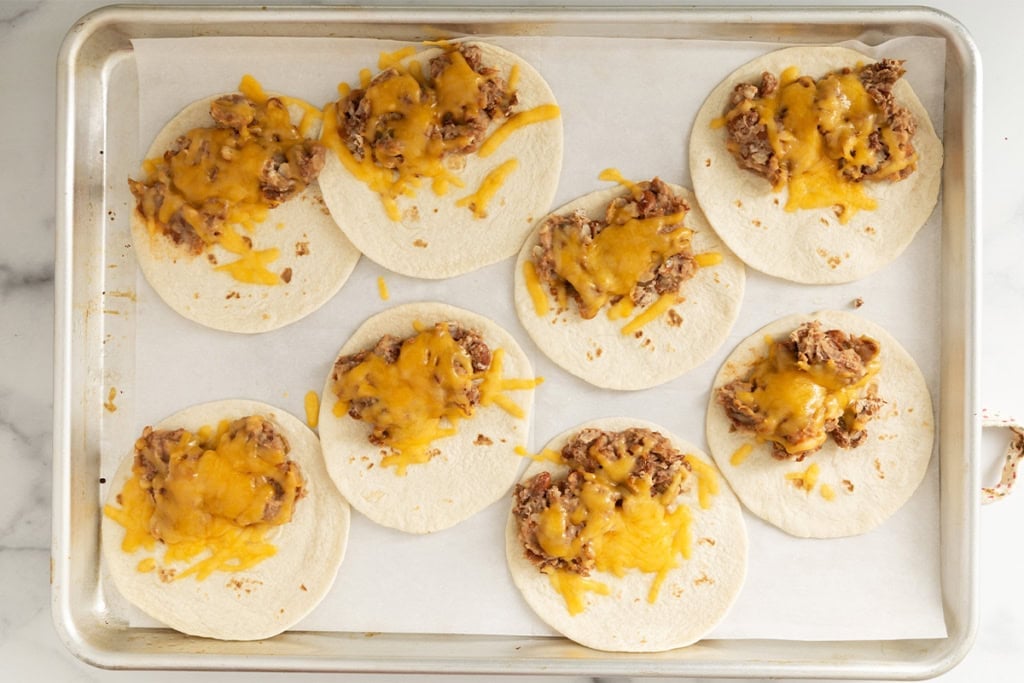 Mini tacos with cheese and beans on baking sheet.