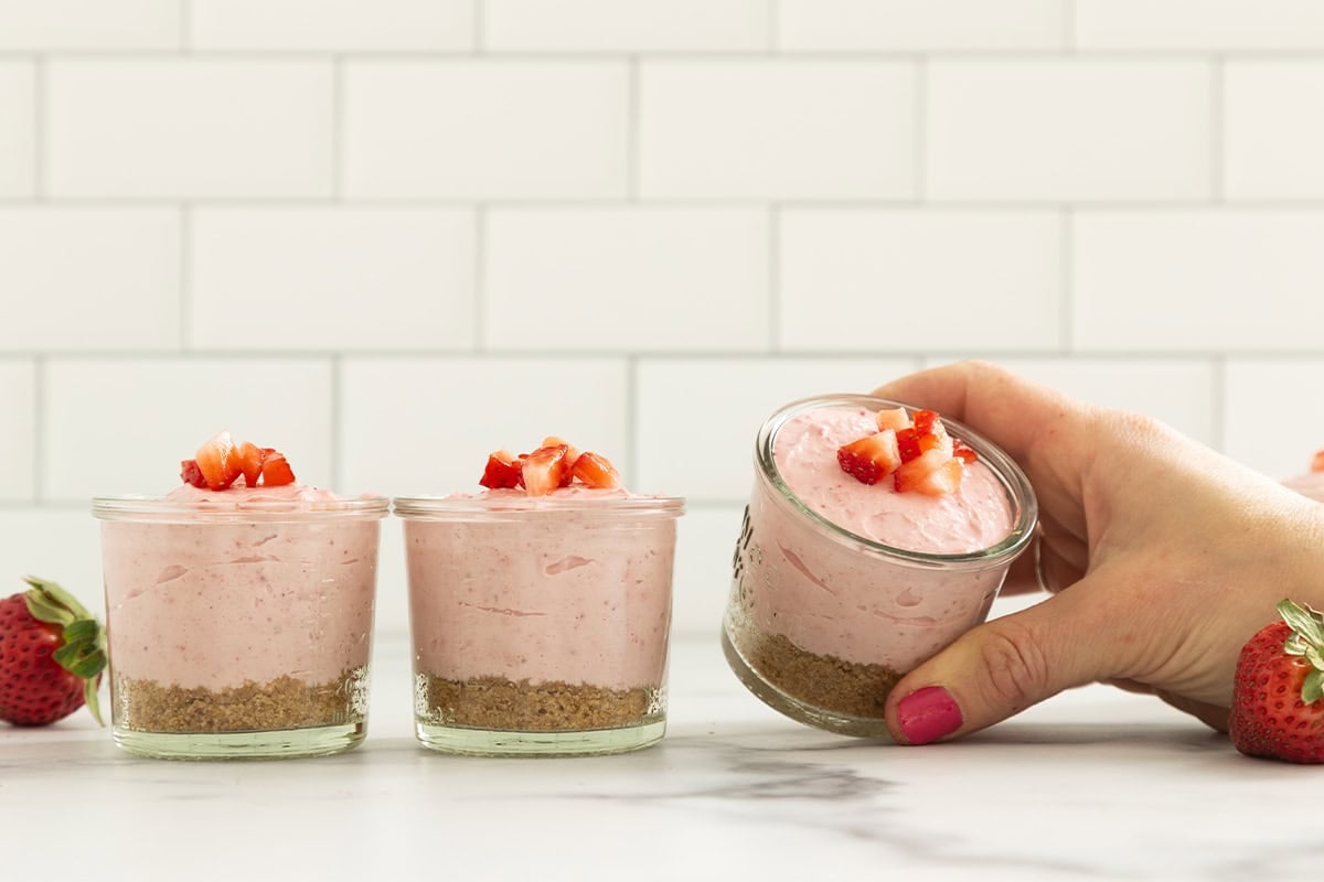 Hands holding glass of no bake strawberry cheesecake.