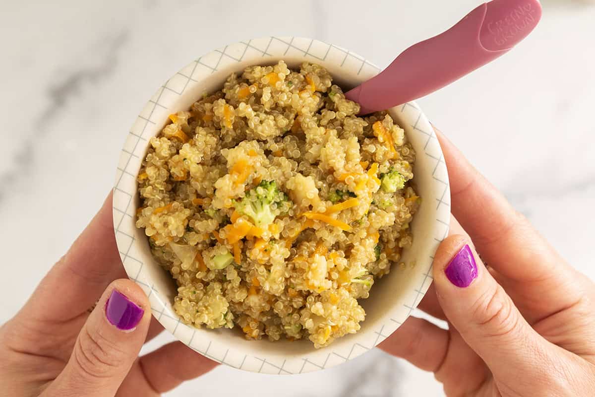 Hands holding bowl of veggie quinoa with pink spoon.