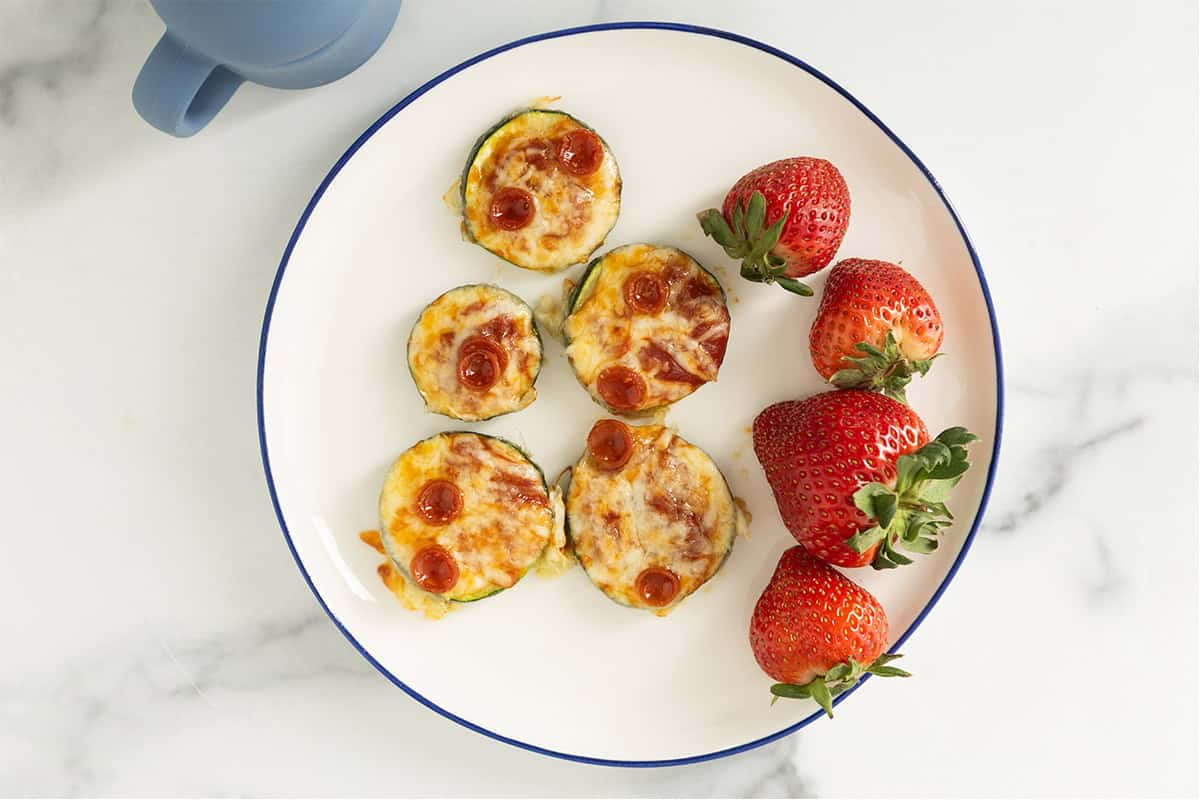 Zucchini pizza bites on white plate with strawberries.