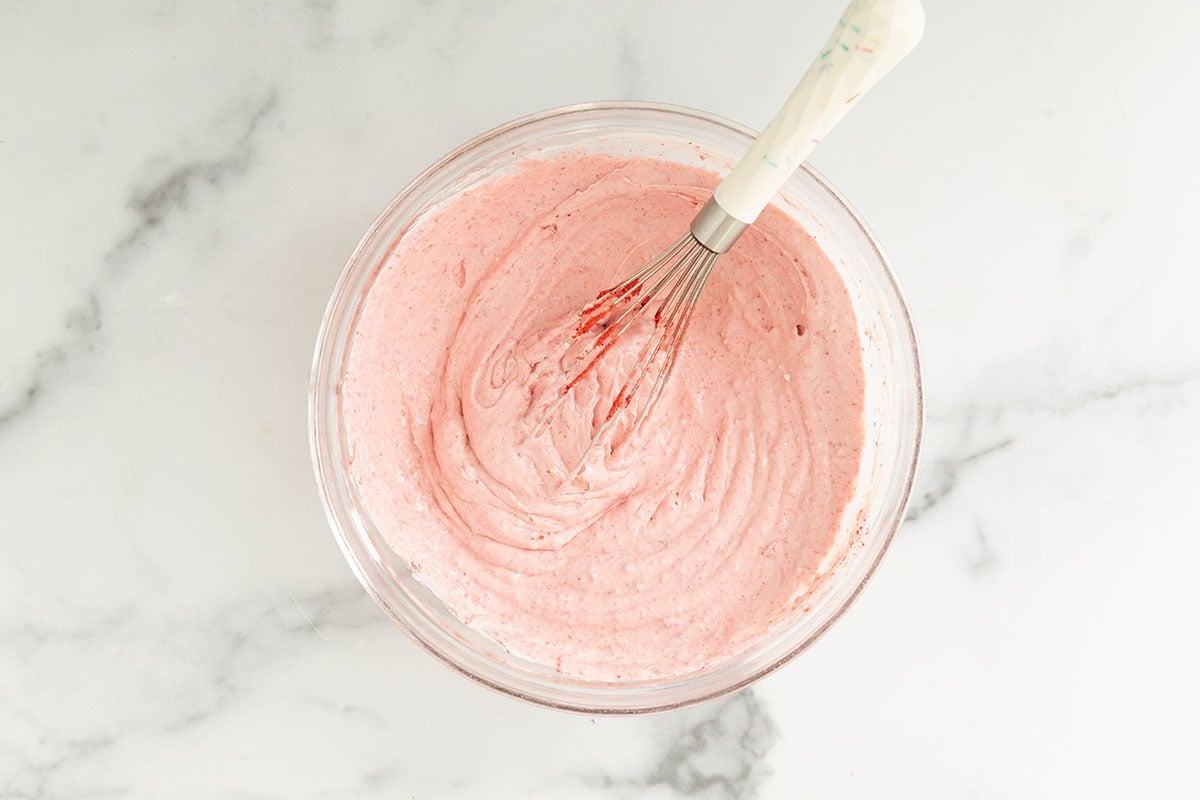Mixture for strawberry cheesecake ice cream in glass bowl with whisk.