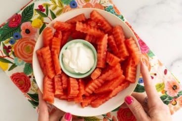 Hands holding plate of watermelon fries with dip in middle.