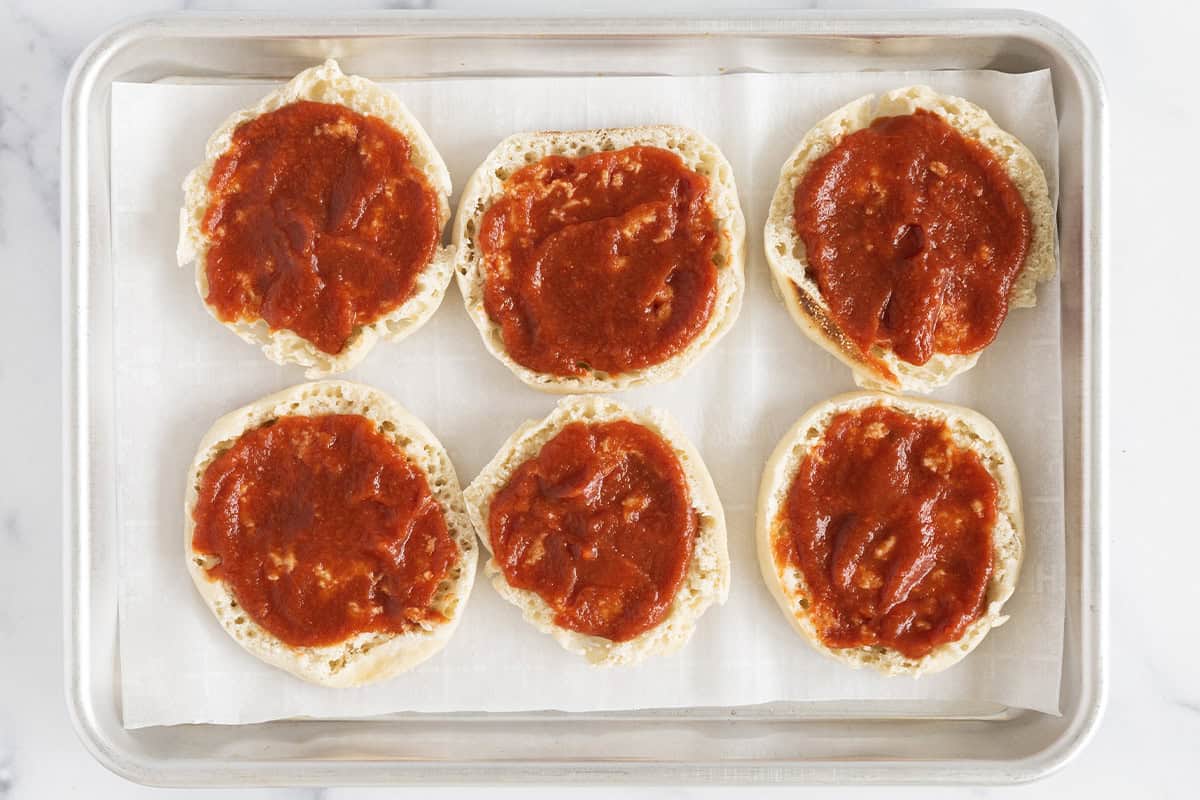 English muffins with pizza sauce on a baking sheet.