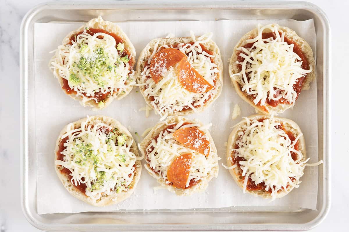 English muffins topped with cheese, pizza toppings, and Parmesan on a baking sheet.