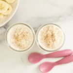 Two jars of banana overnight oats topped with cinnamon beside sliced banana and pink spoons.