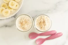 Two jars of banana overnight oats topped with cinnamon beside sliced banana and pink spoons.