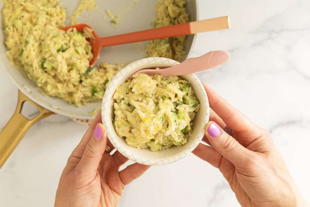 Hands holding a bowl of creamy broccoli orzo with a skillet in the background.
