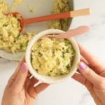 Hands holding a bowl of creamy broccoli orzo with a skillet in the background.